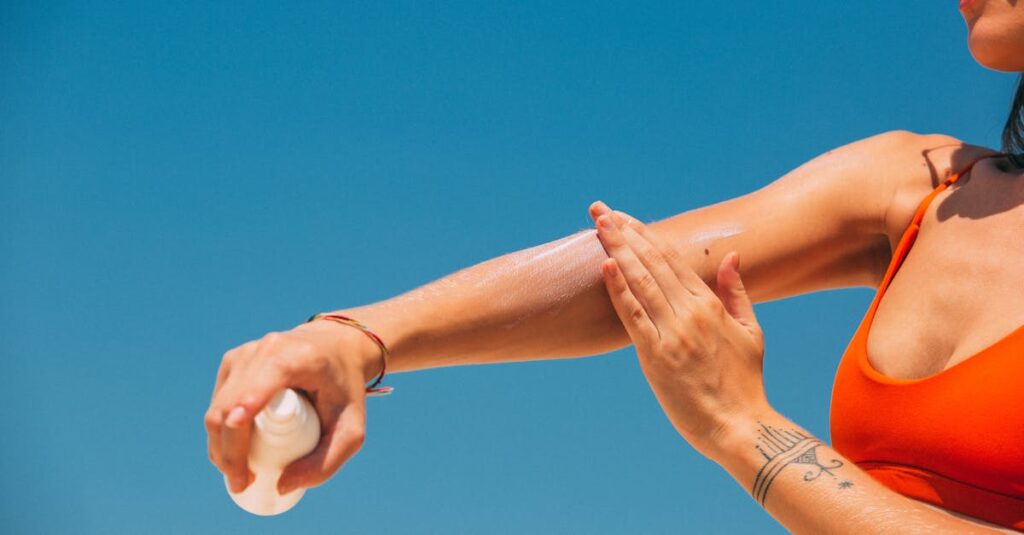 Close-up of a woman applying sunscreen on her arm under a clear blue sky at the beach.